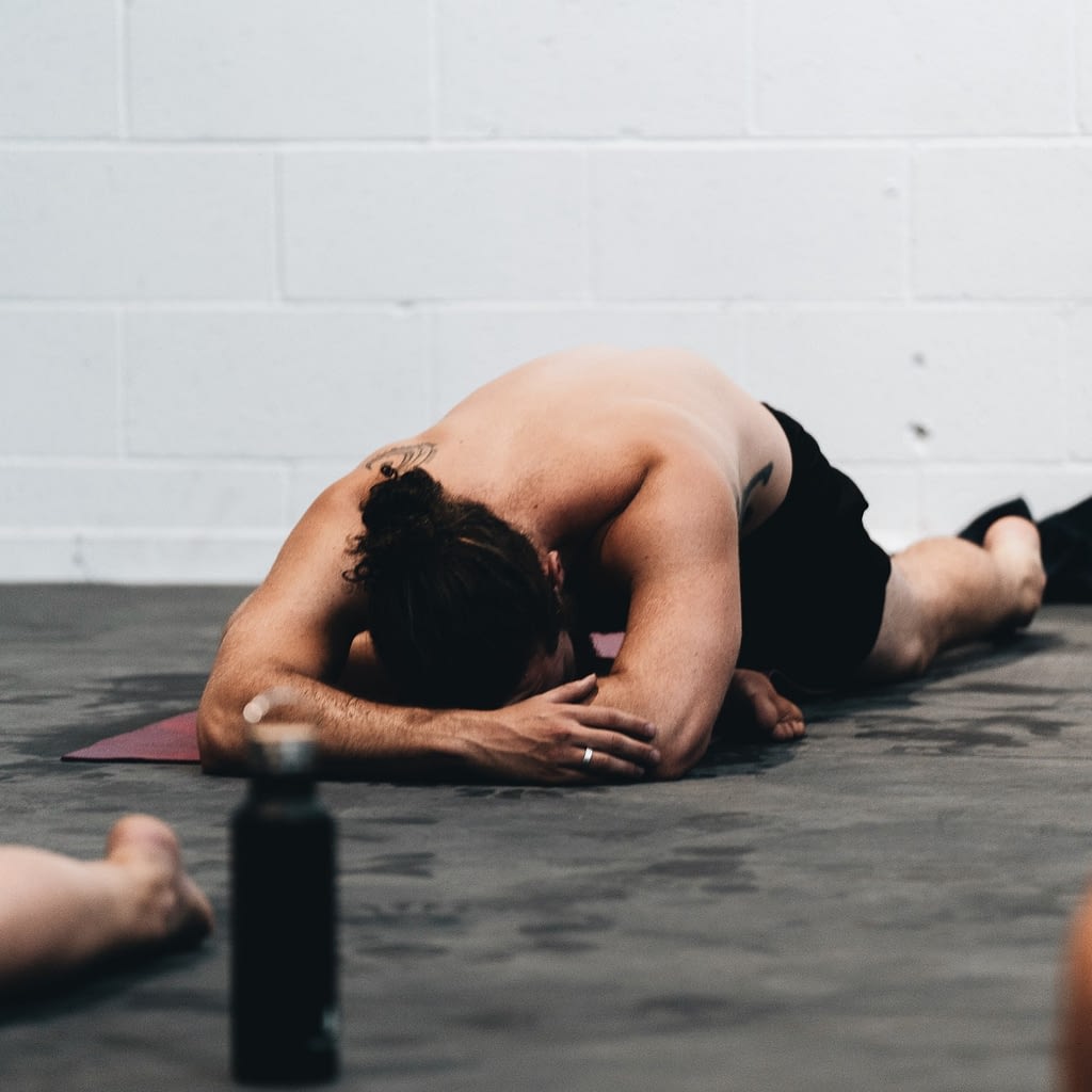 Topless man on exercise mat leaning forward with one foot bent on ground in front and one extended behind, in pigeon pose.