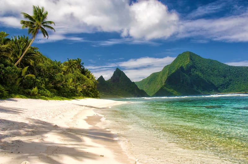 Beach in American Samoa, clear blue-green water lapping against light sand with green mountains in the back
